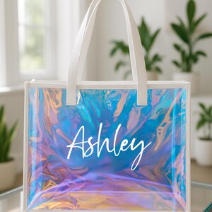 May include: Clear tote bag with white handles and trim. The bag has an iridescent, holographic effect with blue, purple, and gold hues. The name "Ashley" is written in white script across the front.