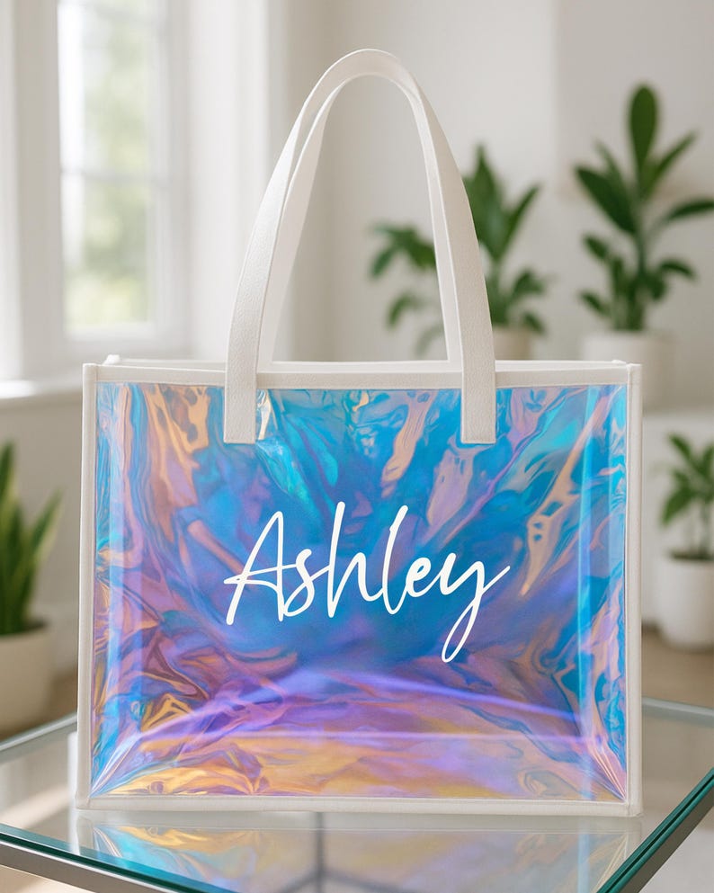 May include: Clear tote bag with white handles and trim. The bag has an iridescent, holographic effect with blue, purple, and gold hues. The name "Ashley" is written in white script across the front.