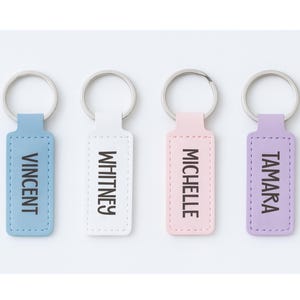 May include: Four personalized keychains in pastel colors: light blue, white, pink, and lavender. Each keychain has a silver ring and a rectangular tag with a name printed in black: Vincent, Whitney, Michelle, and Tamara. The tags have a stitched border.
