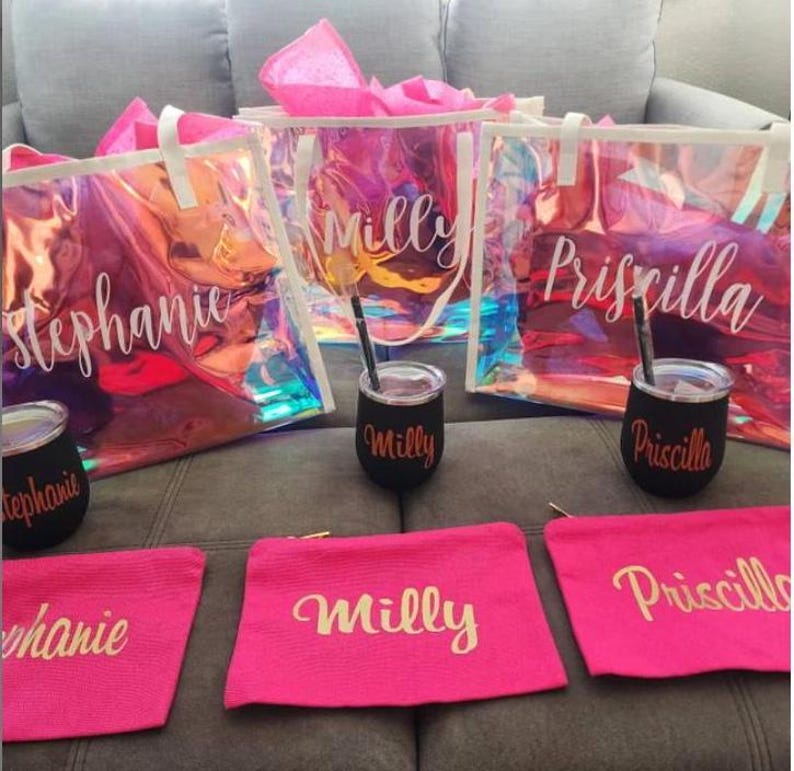 May include: A collection of personalized gifts, including iridescent tote bags, black tumblers with straws, and pink cosmetic bags. Each item is inscribed with a name in a cursive font, creating a cohesive set.