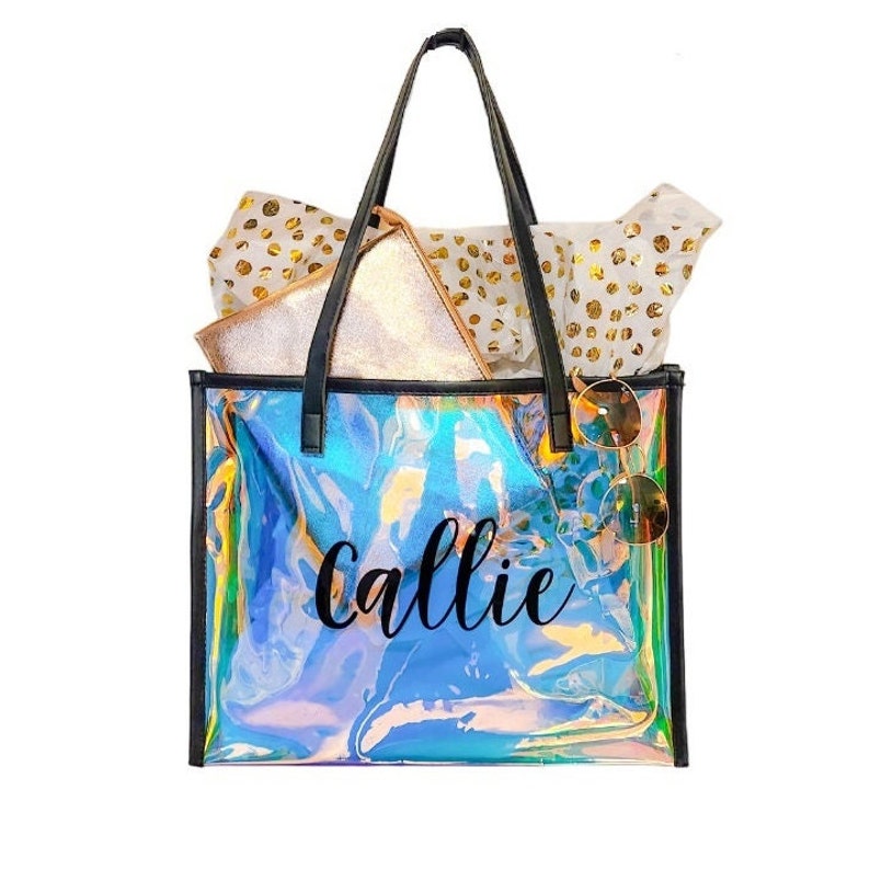 May include: Iridescent tote bag with black trim and handles, featuring the name "callie" in black script. The bag contains a gold clutch, sunglasses, and tissue paper with gold polka dots. A stylish and functional accessory.