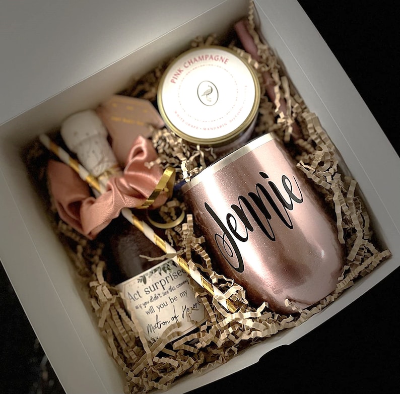 Puede incluir: Una caja de regalo llena de una vela de champ&aacute;n rosado, una mini botella de champ&aacute;n, una pajita, una cinta rosa, un vaso de color rosa dorado personalizado con el nombre "Jennie" y una tarjeta que dice "Act&uacute;a sorprendida si no lo viste venir, &iquest;ser&aacute;s mi dama de honor?"