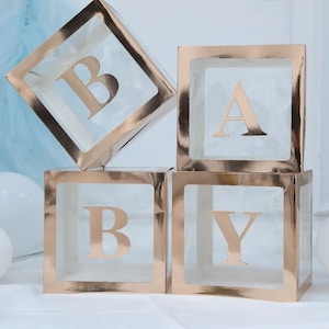 May include: Four clear plastic boxes with gold foil accents, stacked to spell out the word "BABY". Each box has a gold letter inside.