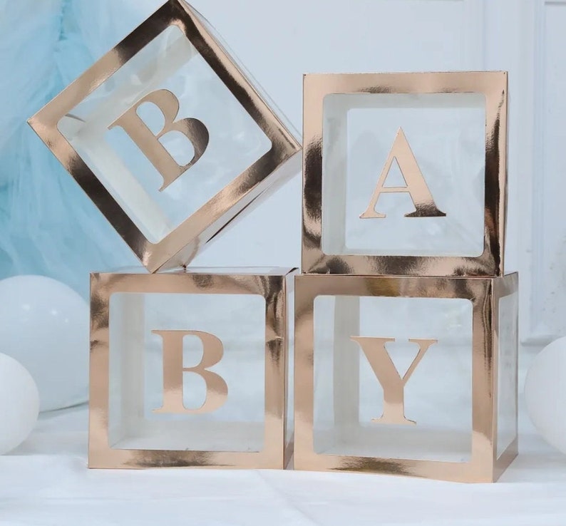 May include: Four clear plastic boxes with gold foil accents, stacked to spell out the word "BABY". Each box has a gold letter inside.