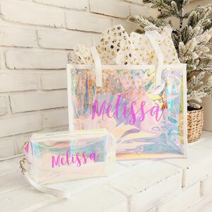 May include: Iridescent tote bag and matching cosmetic bag with the name "Melissa" in pink script. The tote bag has white handles and is filled with gold-speckled tissue paper. The bags sit on a white surface.