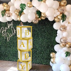 May include: A stack of four clear acrylic boxes with gold trim, each containing white balloons and a gold letter spelling out "BABY". The boxes are in front of a green wall decorated with white and gold balloons.