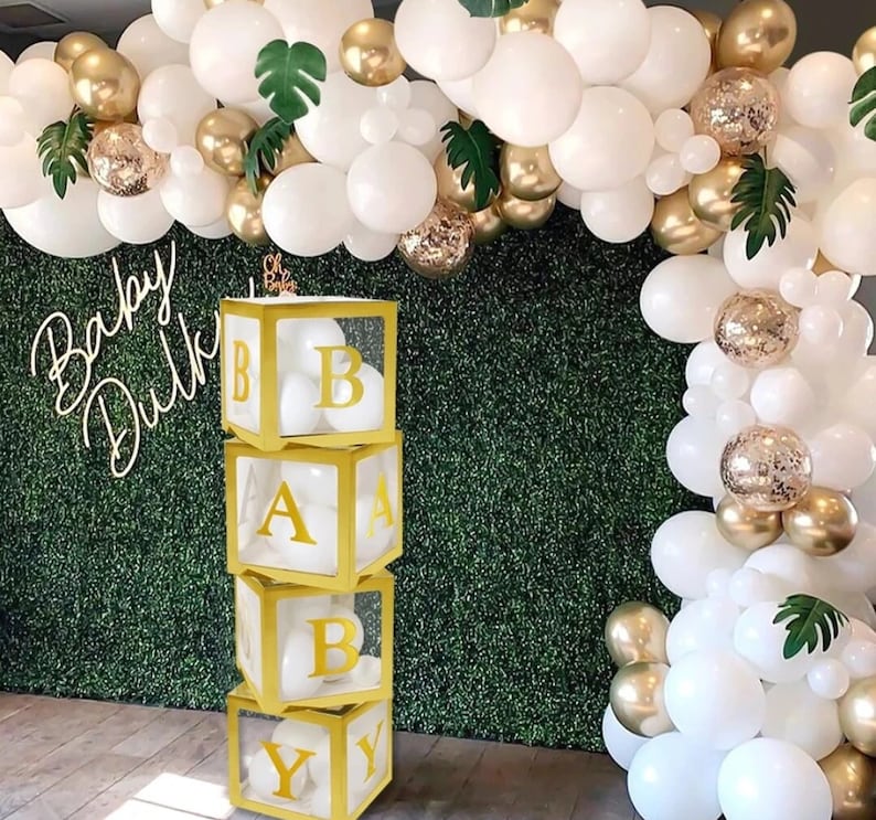 May include: A stack of four clear acrylic boxes with gold trim, each containing white balloons and a gold letter spelling out "BABY". The boxes are in front of a green wall decorated with white and gold balloons.
