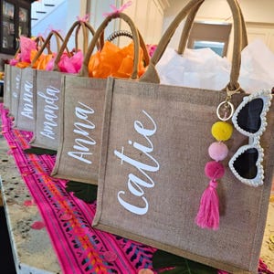 May include: A row of burlap tote bags with white script names. Each bag has a brown handle and is filled with colorful tissue paper. One bag has a keychain with sunglasses and pom-poms.