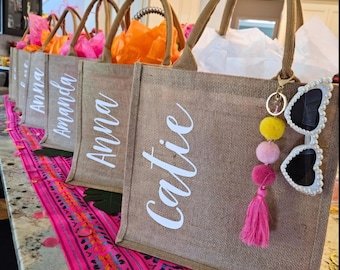 Könnte beinhalten: Eine Reihe von Tragetaschen aus Jute mit weißen Schriftzügen. Jede Tasche hat einen braunen Griff und ist mit farbigem Seidenpapier gefüllt. Eine Tasche hat einen Schlüsselanhänger mit Sonnenbrille und Pompons.