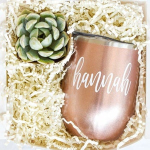 Puede incluir: Una caja de regalo llena de papel blanco triturado, una peque&ntilde;a planta suculenta verde en maceta y un vaso de acero inoxidable color oro rosa con el nombre "hannah" escrito en blanco.