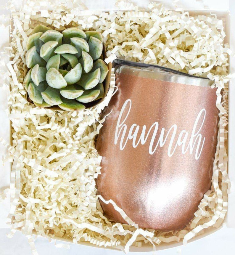 Puede incluir: Una caja de regalo llena de papel blanco triturado, una peque&ntilde;a planta suculenta verde en maceta y un vaso de acero inoxidable color oro rosa con el nombre "hannah" escrito en blanco.
