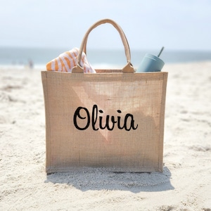 May include: A beige burlap tote bag with the name "Olivia" printed in black cursive. The bag is sitting on a sandy beach and has a striped towel and a blue water bottle inside.