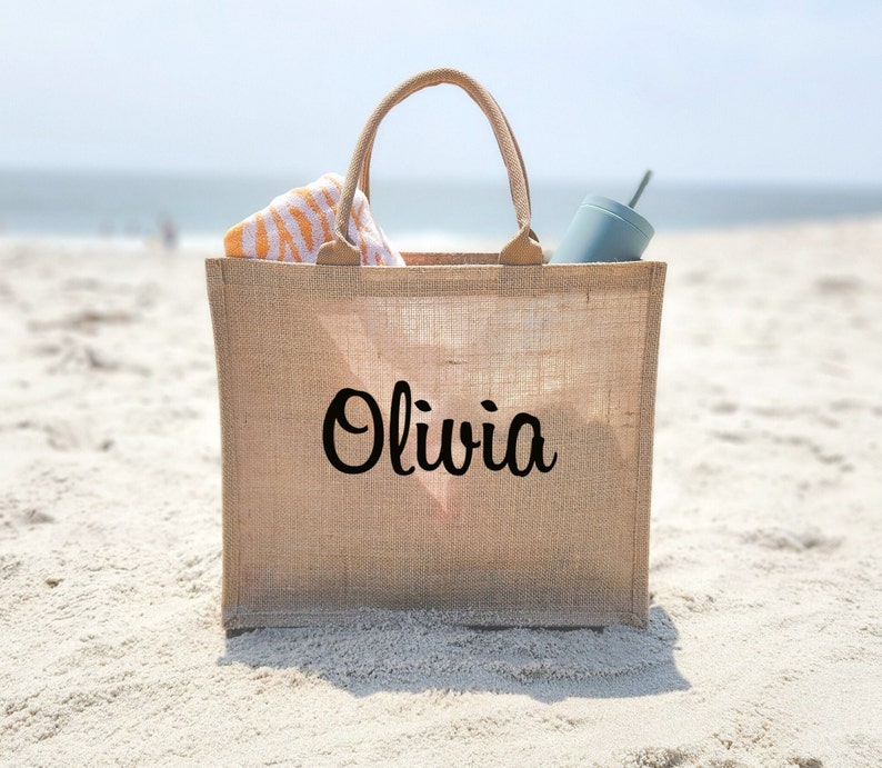 May include: A beige burlap tote bag with the name "Olivia" printed in black cursive. The bag is sitting on a sandy beach and has a striped towel and a blue water bottle inside.