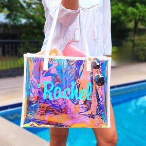 May include: A clear iridescent tote bag with white handles and trim. The bag has the name "Rachel" in turquoise script. Sunglasses hang from the side. The bag contains a pink pouch and a rolled-up towel.