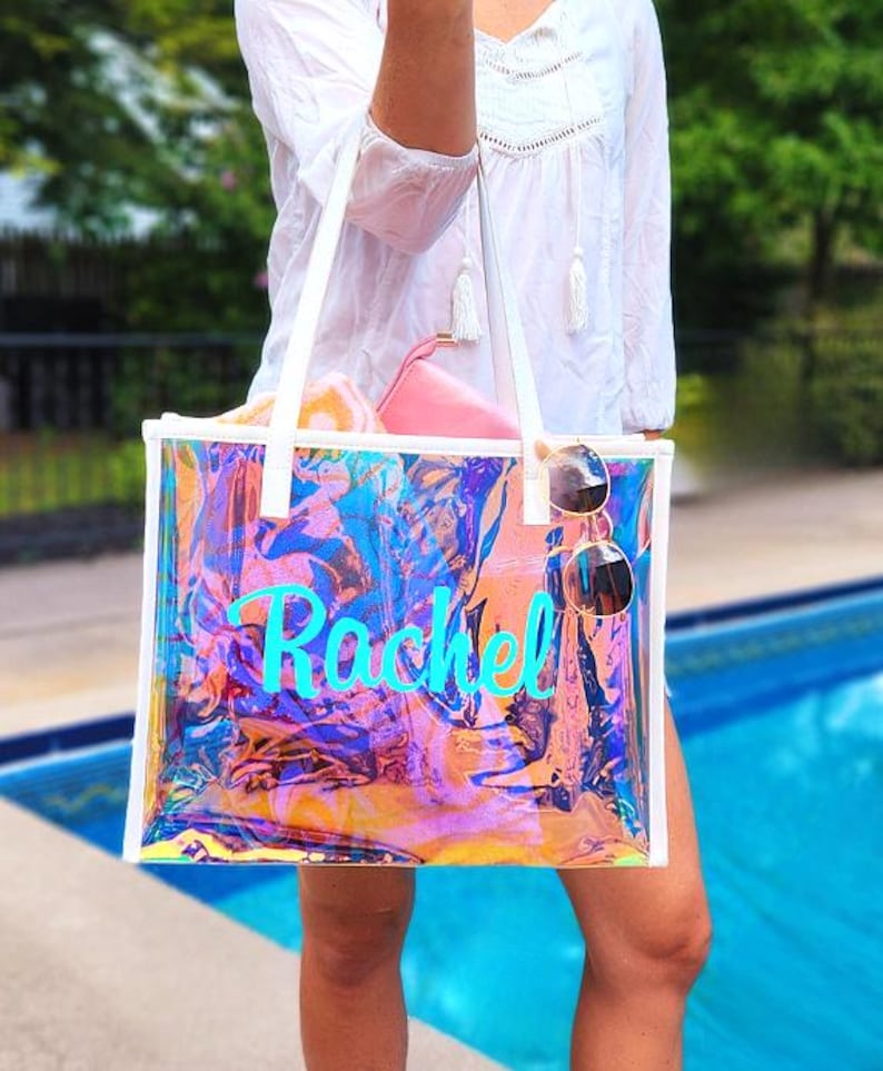 May include: A clear iridescent tote bag with white handles and trim. The bag has the name "Rachel" in turquoise script. Sunglasses hang from the side. The bag contains a pink pouch and a rolled-up towel.