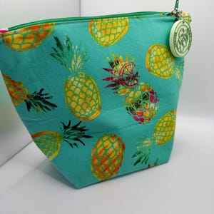 May include: A turquoise fabric cosmetic bag with a repeating pattern of yellow and orange pineapples. The bag has a green zipper and a small, round charm attached to the zipper pull. The bag is designed to stand upright.