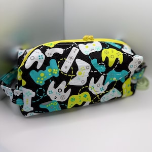 May include: A black zipped pouch featuring a pattern of white, teal, and yellow video game controllers. The pouch has a yellow zip and a teal and black handle. The background is white.