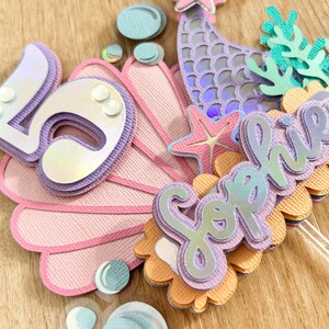 May include: A mermaid-themed cake topper with a pink seashell, a purple mermaid tail, a starfish, and the name "Sophia" in purple letters.