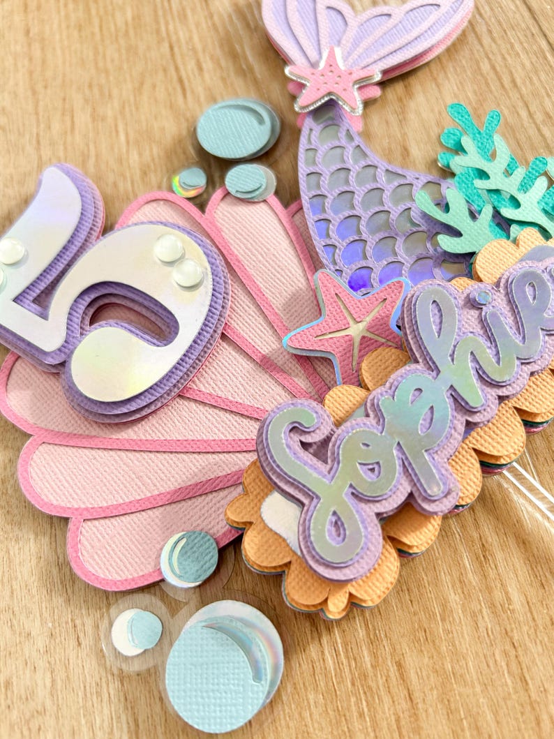 May include: A mermaid-themed cake topper with a pink seashell, a purple mermaid tail, a starfish, and the name "Sophia" in purple letters.