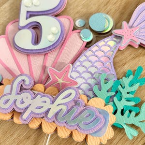 May include: A mermaid-themed cake topper with a purple and white number 5, a purple mermaid tail, a pink seashell, a starfish, and the name "Sophie" in purple letters.