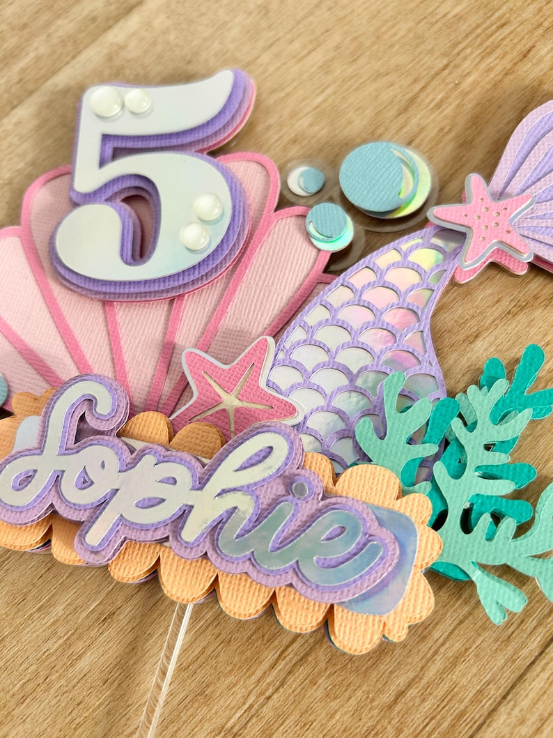 May include: A mermaid-themed cake topper with a purple and white number 5, a purple mermaid tail, a pink seashell, a starfish, and the name "Sophie" in purple letters.