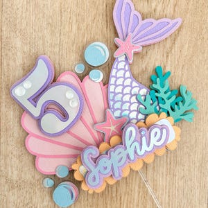 May include: A mermaid themed cake topper with a purple mermaid tail, pink seashells, a starfish, and custom name in silver letters. The topper is on a cardstock background.