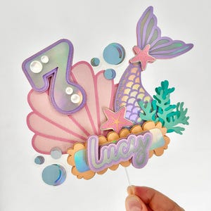May include: A mermaid themed cake topper with a purple mermaid tail, a pink seashell, a light blue bubble, a starfish, and the name 'Lucy' in purple and orange letters. The number '7' is also included in the topper.