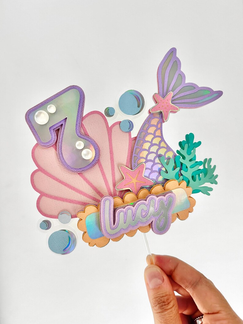 May include: A mermaid themed cake topper with a purple mermaid tail, a pink seashell, a light blue bubble, a starfish, and the name 'Lucy' in purple and orange letters. The number '7' is also included in the topper.