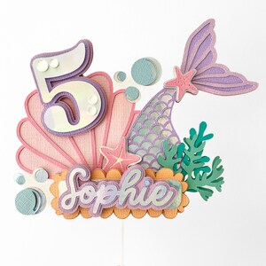 May include: A mermaid-themed cake topper with a pink seashell, a purple mermaid tail, a number 5, and the name "Sophie" in iridescent lettering.