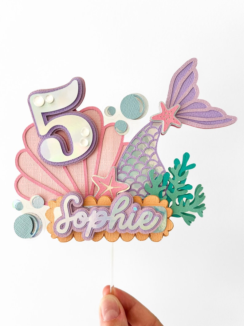 May include: A mermaid-themed cake topper with a pink seashell, a purple mermaid tail, a number 5, and the name "Sophie" in iridescent lettering.