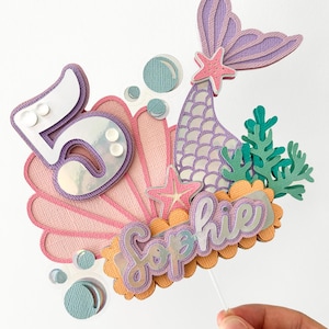 May include: A mermaid-themed cake topper with a purple and pink seashell, a purple mermaid tail, a starfish, and the name "Sophie" in purple and orange lettering. The number "5" is also included in the design.