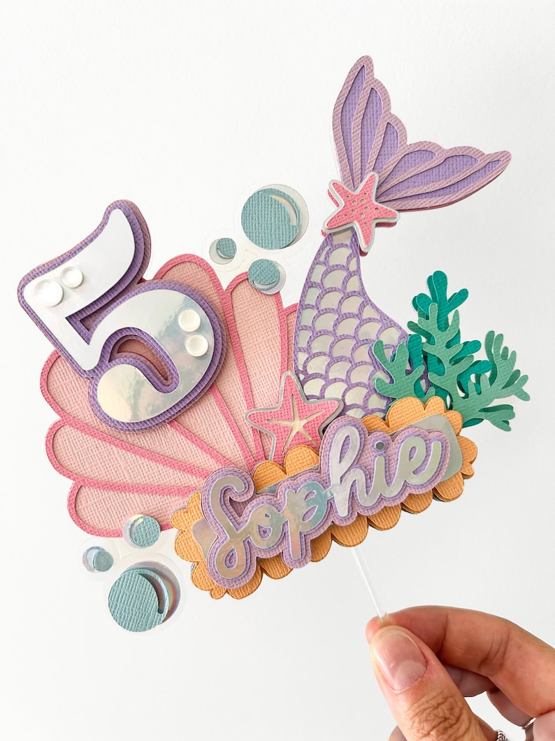 May include: A mermaid-themed cake topper with a purple and pink seashell, a purple mermaid tail, a starfish, and the name "Sophie" in purple and orange lettering. The number "5" is also included in the design.