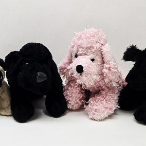 Op de afbeelding: Vier pluche honden. Een beige mopshond met zwarte accenten, twee zwarte honden en een roze poedel. De knuffels zijn zacht en pluizig, met gedetailleerde ogen en neuzen. De honden zitten rechtop.