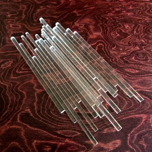 May include: A group of clear acrylic rods of varying lengths, arranged in a fan shape on a dark wood surface.