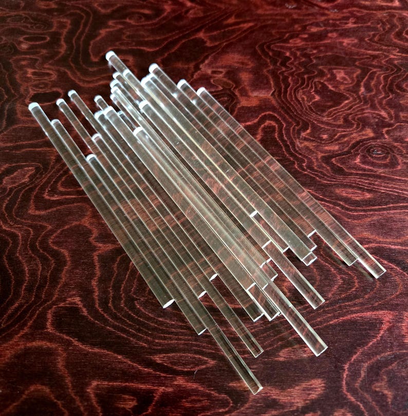 May include: A group of clear acrylic rods of varying lengths, arranged in a fan shape on a dark wood surface.