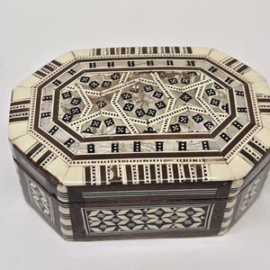 May include: An octagonal wooden jewelry box with intricate inlaid mother-of-pearl and dark wood geometric patterns. The box features a cream-colored border and a detailed design on the lid and sides, showcasing craftsmanship.