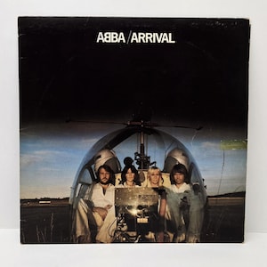 Puede incluir: Portada del álbum "Arrival" de ABBA. La imagen presenta a los miembros de la banda dentro de un helicóptero. El título del álbum está impreso en blanco en la parte superior. El fondo es un cielo oscuro con un campo.