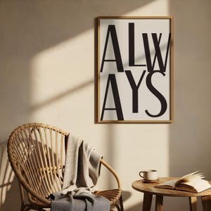 Puede incluir: Un cuadro enmarcado con la palabra "ALWAYS" en letras negras sobre un fondo crema. La impresión está en un marco de madera marrón claro. Una silla de mimbre con una manta y una pequeña mesa de madera con una taza y un libro están en primer plano.