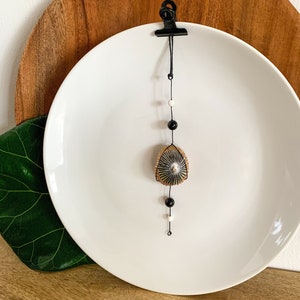 May include: A decorative hanging ornament featuring a triangular, woven design with a silver center, suspended from a black clip. The ornament is adorned with black and white beads, displayed against a white plate and wooden background.