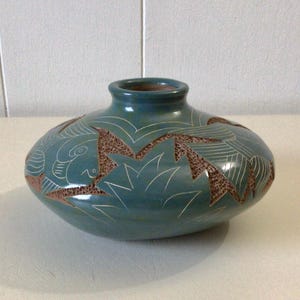 May include: A teal-colored ceramic vase with a rounded body and a narrow neck. The vase features intricate carved designs in a contrasting brown color, including geometric shapes and stylized patterns. The vase is a decorative item.