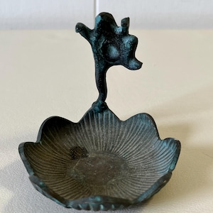 May include: A small, antique-style bronze dish with a stylized figure on a stem. The dish has a scalloped edge and a textured, sunburst pattern. The figure is dark teal with a weathered appearance.