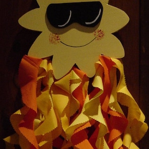 May include: A yellow sun with a smiling face and sunglasses, hanging from a string. The sun is surrounded by a cascade of orange and yellow fabric.