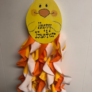 May include: A yellow chick-shaped wooden sign with the words "Happy Easter" painted on it. The sign is hanging from a white string and has a white, orange, and yellow fabric garland attached to the bottom.