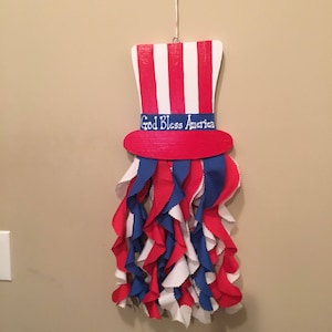 May include: A red, white, and blue Uncle Sam hat with the words "God Bless America" on the band. The hat is hanging from a white string and has red, white, and blue fabric streamers hanging from the bottom.