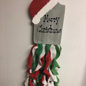May include: A wooden sign with a red and white Santa hat on top. The sign is painted gray and has the words "Merry Christmas" written in black. A long strand of red, white, and green fabric hangs from the bottom of the sign.