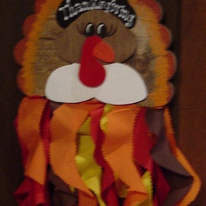 May include: A brown and orange felt turkey with a black hat that says "Happy Thanksgiving". The turkey has a white beard and a red wattle. The turkey is decorated with brown, orange, red, and yellow felt feathers.