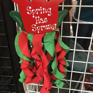May include: A red wooden tulip with green leaves and the text "Spring Has Sprung" hanging from a white wire rack. The tulip has red and green fabric streamers hanging from it.