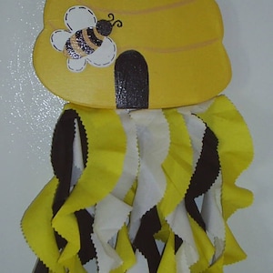 May include: A yellow and black beehive-shaped welcome sign with a painted bee and the word "Welcome". The sign is hanging from a white string and has a fringe of yellow, white, and black fabric.