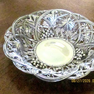 May include: A silver-toned metal bowl featuring an intricate openwork border decorated with embossed grape clusters and vine leaves. The center of the bowl is smooth and circular, contrasting with the detailed, textured filigree rim.