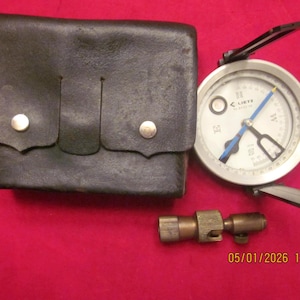 May include: A vintage compass with a black leather case and a brass fitting. The compass face is white with black and blue markings. The date 05/01/2026 18:52 is printed in the lower right corner. The case is closed with two silver snaps.